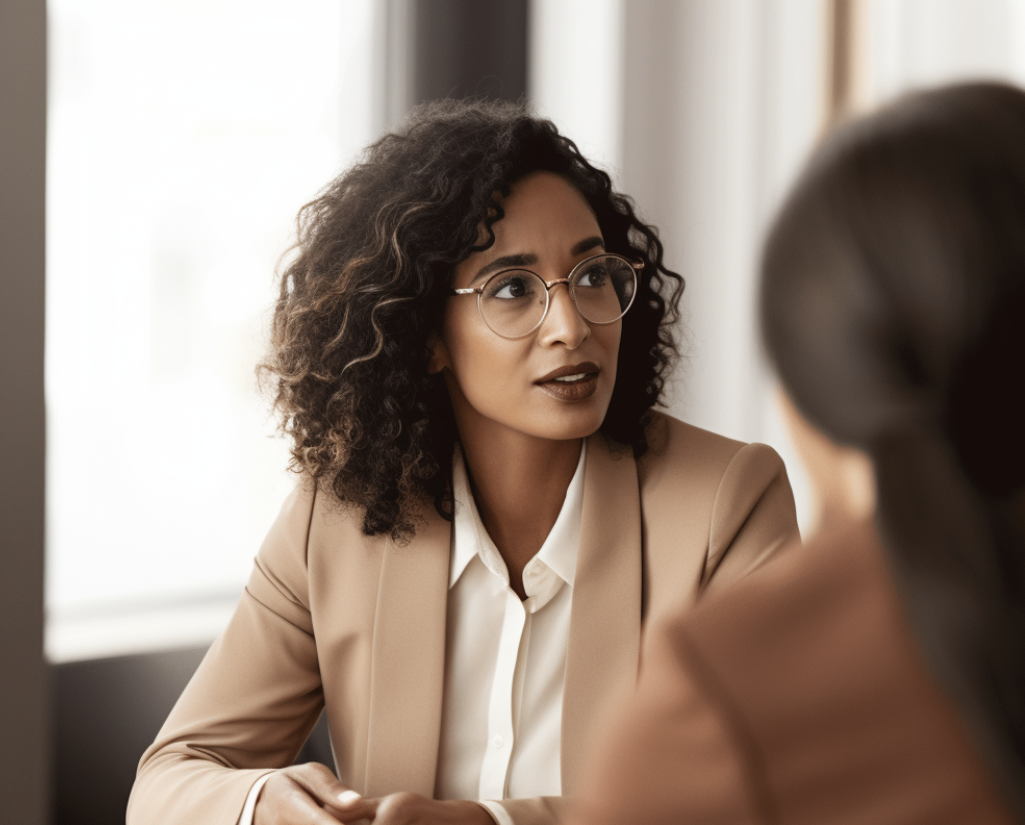 An attorney with curly hair and glasses, dressed in a beige blazer and white blouse, is seated across from someone, engaged in conversation.